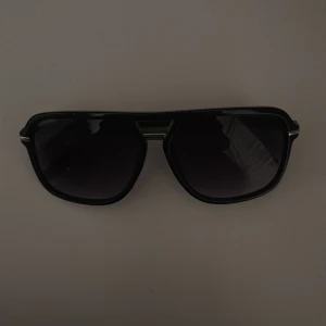 Svarta pilotbågar solglasögon - Stilrena svarta solglasögon med klassisk pilotform och mörka glas. Bågarna är i plast med en metallbrygga över näsan för extra detalj. Perfekta för en cool look i solen.