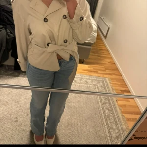 Beige trenchcoat med knytbälte - Snygg beige trenchcoat med dubbelknäppning och knytbälte i midjan. Jackan är använd fåtal gånger 