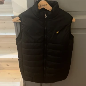 Svart dunväst från Lyle & Scott - Snygg svart dunväst från Lyle & Scott med dragkedja och en liten gul logga på bröstet. Perfekt för lager-på-lager under kyliga dagar. Västen har en stilren design och är lätt att matcha med olika outfits.