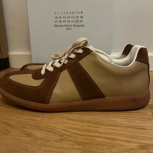 Bruna och beige sneakers från Maison Martin Margiela - Snygga sneakers i en stilren design från Maison Martin Margiela. Skorna har en kombination av brunt och beige med snörning framtill. Perfekta för en avslappnad men trendig look.