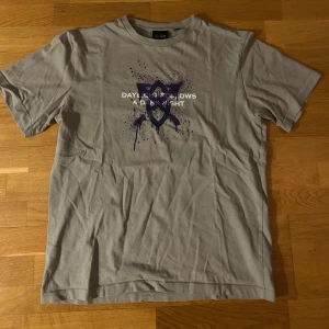 Grå t-shirt från Daily Paper - Säljer en grå t-shirt från Daily Paper med ett coolt tryck i lila och texten 'DAYLIGHT FOLLOWS A DARK KNIGHT'. Perfekt för en avslappnad stil. Passar både M och L