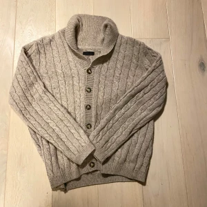 Beige kabel stickad cardigan - Mysig beige stickad kofta från Cebus med kabelstickat mönster och knappar framtill. Perfekt för kyliga dagar med sin varma och bekväma design. I bra skick