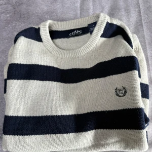 Randig tröja från Chaps - Säljer en snygg randig tröja från Chaps i marinblått och vitt. Tröjan har långa ärmar och ett broderat emblem på bröstet. Perfekt för en stilren look!