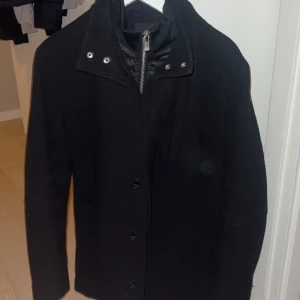 Svart rock med dragkedja och knappar - Snygg svart rock med både dragkedja och knappar framtill. Jackan har en stilren design med långa ärmar och hög krage. Perfekt för kyligare dagar. Jackan är från Jack and Jones och köptes för 2499kr passar M samt S sitter perfekt i nederdelen