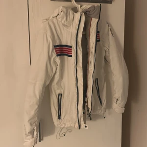 Vit jacka från Helly Hansen - Säljer en vit jacka från Helly Hansen med coola ränder i rött och blått på bröstet och ryggen. Jackan har en praktisk dragkedja och huva, perfekt för kyliga dagar. Den har en normal passform och är tillverkad i slitstarkt material.