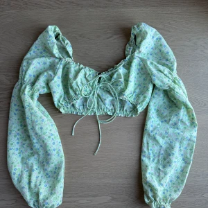 Grön blommig croppad blus - Söt grön blus med blommigt mönster i blått och rosa. Blusen är croppad med långa ärmar och har en knytning framtill. Perfekt för en somrig look!