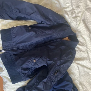 Mörkblå bomberjacka från Alpha Industries - Säljer en snygg mörkblå bomberjacka från Alpha Industries. Jackan har en klassisk design med dragkedja och ribbade muddar. Jackan är i super bra skick. Perfekt för en cool och klassisk Sthlms stil. Passar både tjej och kille.