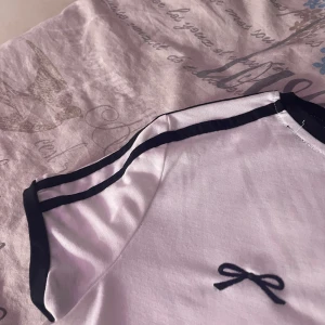 Vit kortärmad topp med svart rosett - Säljer en söt vit topp med korta ärmar och svarta detaljer. Toppen har en liten svart rosett på bröstet och svarta ränder längs ärmarna. Perfekt för en avslappnad stil.