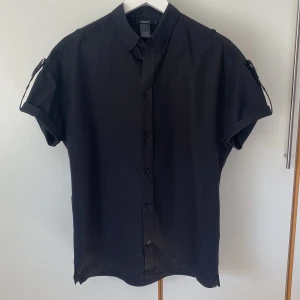 Svart kortärmad skjorta från Filippa K - Cool svart kortärmad skjorta från Filippa K. Skjortan har knappar framtill och uppvikbara ärmar i flera steg. knappdetaljer på ärmarna. Från Filippa K Man men är lika snygg på en tjej. 