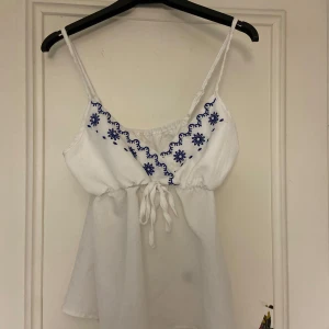Vit linne med blå broderier - Charmig vit linne med smala axelband och vackra blå broderier längs halsringningen. Toppen har en knytning framtill och en luftig passform, perfekt för varma dagar.