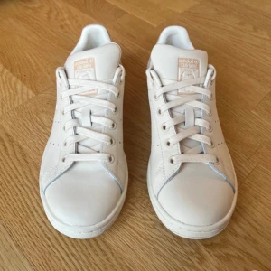 Begie/krämvita Adidas Stan Smith sneakers - Snygga vita Adidas Stan Smith sneakers med klassisk design. Skorna har en ljusrosa detalj på hälen och perforerade ränder på sidorna. Perfekta för en stilren look.