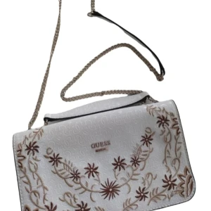 Vit broderad axelväska från Guess - Snygg vit axelväska från Guess med vackra broderade blommor i beige och brunt. Väskan har en elegant kedjerem och subtilt mönster i tyget. Perfekt för att ge en stilfull touch till din outfit. (Som ny, bara etiketten som fattas)