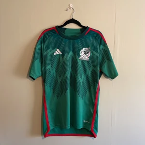 Mexico fotbollströja från Adidas - Säljer en grön fotbollströja från Adidas med Mexikos landslagsemblem på bröstet. Tröjan har ett snyggt mönster i mörkare grönt och röda detaljer längs sidorna och axlarna. Perfekt för fotbollsfans som vill visa sitt stöd för Mexiko! 💚