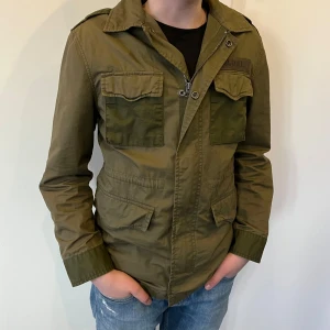 Olivgrön jacka från Polo Ralph Lauren - Säljer en olivgrön jacka från Polo Ralph Lauren med dragkedja och knappar framtill. Jackan har flera praktiska fickor och en klassisk krage. Perfekt för höst och vår! Priset kan diskuteras också.😁