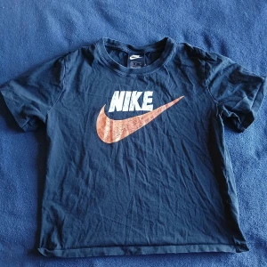 Svart t-shirt från Nike - Säljer en svart t-shirt från Nike med det klassiska logotyptrycket i vitt och orange på framsidan. Perfekt för en sportig look eller avslappnad stil. Tillverkad i mjukt material för bekväm passform.