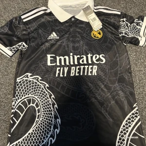 Real Madrid fotbolls T-shirt  - Snygg svart fotbollströja från Adidas med ett coolt drakmönster. Tröjan har en vit krage och vita detaljer på ärmarna. Den har korta ärmar och klubbens emblem på bröstet. Perfekt för fotbollsfans som vill sticka ut!