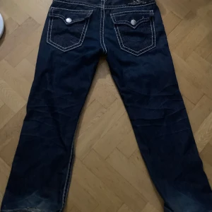 True Religion Jeans - En del av stitchingen på ena bakfickan är trasig Snygga mörkblå jeans från True Religion med kontrasterande sömmar och klassisk femficksdesign. Tillverkade i USA, dessa jeans har en stilren look med knappar och dragkedja framtill. Perfekta för en avslappnad stil.