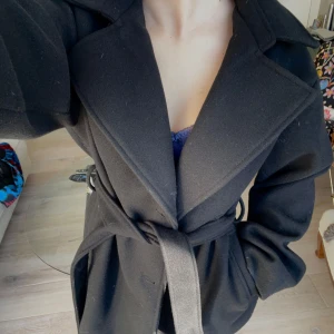 Kort kappa från NA-KD - Snygg svart kort kappa från NA-KD med knytskärp i midjan. Den har en klassisk design med breda slag och långa ärmar. Köpt på NA-KD för 899kr och har bara använts två gånger!🥳