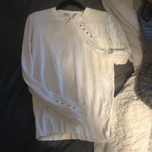 Vit långärmad tröja från Zara - Säljer en stilren vit långärmad tröja från Zara i mjukt material. Tröjan har rund hals och snygga knappar på ärmsluten som ger en extra detalj. Storlek M