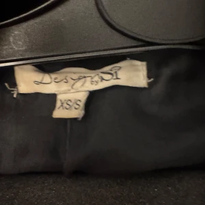 Svart skepparkavaj från Desigual XS/S - Klassisk svart skepparkavaj från Desigual i storlek XS/S. Jackan har breda slag, dubbelknäppning med svarta knappar och två fickor framtill. Perfekt för dig som gillar en tidlös och stilren look med lite edge.