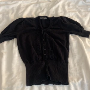 Svart kofta från Miss Ewing - Svart kofta från Miss Ewing i storlek 40 men den är som XS/S. Den har rund hals, knappar hela vägen framtill och snygga stickade detaljer längs bröstet. Koftan har puffade trekvartsärmar och bred ribbad kant nedtill som ger en cool siluett. 