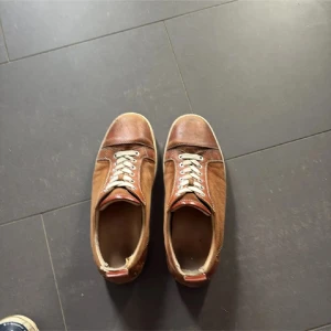 Bruna sneakers med snörning - Säljer ett par bruna sneakers med vit snörning och rund tå. Skorna har en klassisk låg profil och är tillverkade i skinn med detaljer i olika bruna nyanser. Perfekta för dig som gillar retrostil och vill ha något som sticker ut.