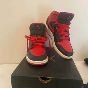 Nike Jordan 1 mid röd/svart EUR 39 - Nike Air Jordan 1 midi rött och svart med klassisk Jumpman-logga på plösen. Skorna har snörning, rund tå och vit platt sula. Ovandelen är i syntet och skinn, med röda och svarta paneler som ger en riktigt fet kontrast. Perfekt för dig som vill sticka ut med dina sneakers.