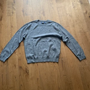 Grå stickad tröja från Polo Ralph Lauren - Säljer en grå stickad tröja från Polo Ralph Lauren i storlek M. Tröjan har rund hals, långärmade och ett diskret broderat Polo-logo på bröstet. Mjuk och skön kvalitet som passar perfekt till jeans eller chinos. Klassisk och stilren look.