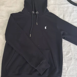 Svart hoodie från Polo Ralph Lauren - Svart hoodie från Polo Ralph Lauren med klassisk broderad logga i vitt på bröstet. Tröjan har huva med snörning, stor magficka och långa ärmar. Mjuk och skön bomullskvalitet, perfekt för chill dagar. Snygg och enkel design som funkar till allt.
