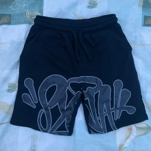 Svarta shorts Syna med tryck och snörning - Oanvända! Svarta shorts från Syna i 100% bomull med stort grått tryck framtill i graffiti-stil. De har elastisk midja med snörning och praktiska fickor med dragkedja. Perfekta för chill dagar eller när du vill ha en streetwear vibe.