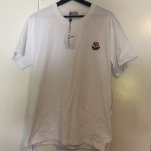 Vit Moncler t-shirt med logga - Vit t-shirt från Moncler i mjuk bomull med rund hals och korta ärmar. På bröstet sitter en snygg broderad Moncler-logga i blått och rött. Enkel och clean design som funkar till allt. Perfekt för dig som gillar stilrena märkesplagg.