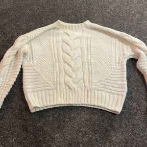 Vit stickad tröja med flätmönster - Säljer en vit stickad tröja från H&M Divided i storlek XS. Tröjan har ett snyggt flätmönster framtill och längs ärmarna, samt ribbade muddar vid hals, ärmslut och nederkant. Perfekt för dig som gillar chunky knit och vill ha en mysig look.