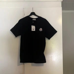 Svart Moncler t-shirt med logga - Svart t-shirt från Moncler med klassisk rund hals och korta ärmar. På bröstet sitter en snygg broderad Moncler-logga i vitt, blått och rött. Materialet är mjuk bomull som känns skön mot huden. Perfekt för dig som gillar stilrena designer och exklusiva märken.