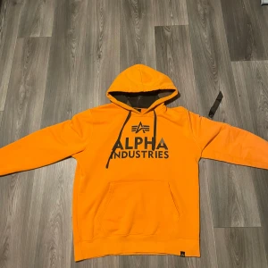 Orange hoodie från Alpha Industries - Säljer en riktigt snygg orange hoodie från Alpha Industries i storlek L. Tröjan har stor logga framtill, och en klassisk magficka. Perfekt för dig som gillar streetwear och vill sticka ut med färg. Mjuk insida och skön passform.