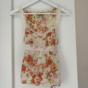 Blommig linnetopp från MNG  - Supersöt linnetopp från MNG Suit i storlek S, med romantiskt blommigt mönster i rosa, orange och grönt på en ljusbeige botten. Toppen har spetsdetaljer vid axelband och midja samt knytband i ryggen. Perfekt för sommarens chill dagar.