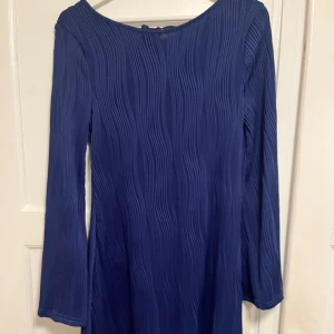 Marinblå långärmad klänning med knyt - Snygg marinblå klänning med lång ärm och vågigt strukturerat tyg. Klänningen har en djup, rundad rygg med knytband upptill för extra detalj. Passformen är normal och tyget känns mjukt och följsamt. Perfekt för dig som gillar stilrena plagg med twist.