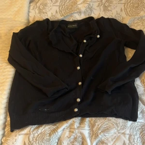 Svart kofta med pärlknappar från Anna Field - Svart kofta från Anna Field med snygga pärlknappar framtill. Tröjan har långärmad design och är tillverkad i mjuk viskosblandning med polyamid. Perfekt för dig som gillar klassisk stil med en twist. Passar dig som vill ha en enkel men elegant look. Har lite smuts som går bort i tvätten, annars jättebra skick och har inga defekter 💕