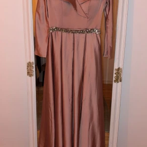 Lång rosa klänning med paljettbälte - Säljer en elegant långklänning i mjukt rosa tyg med glansig finish. Klänningen har långa ärmar, v-ringad omlottkrage och ett snyggt paljettbälte i midjan som ger extra bling. Perfekt för bal eller fest! Materialet känns lyxigt och faller snyggt.