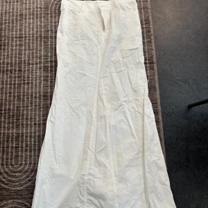 Vit långkjol med utsvängd form - Säljer en vit långkjol med utsvängd siluett och stilrena sömmar framtill. Kjolen har hällor för bälte och dragkedja i midjan. Perfekt för dig som gillar minimalistisk och clean stil.