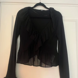 Svart volangblus från Amisu - Säljer en svart blus från Amisu i storlek M. Blusen har långa ärmar, v-ringning med volangkant och en snygg knytning framtill. Nederdelen är lätt transparent med volangdetaljer som ger en extra touch. Perfekt för dig som gillar romantiska vibes och coola detaljer.