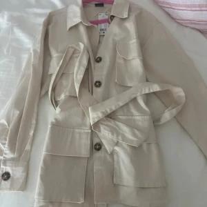 Beige overshirt med bälte och fickor - Stilren beige overshirt från Gina Tricot med stora fickor framtill, knappar och matchande bälte i midjan. Långärmad modell i bomull, perfekt för lager-på-lager och en clean look.