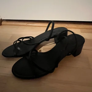 Svarta skor med klack - Säljer ett par svarta skor med låg klack och tunna remmar som korsar över foten. Köpta på H&M och använda en gång!