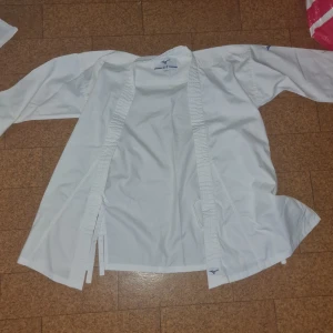 Vit karatekostym från Mizuno - Säljer en klassisk vit karatekostym från Mizuno med tillhörande bälte. Kostymen består av en långärmad jacka med förstärkta sömmar och ett par raka byxor med resår i midjan. Materialet är slitstark bomull, perfekt för träning och tävling.