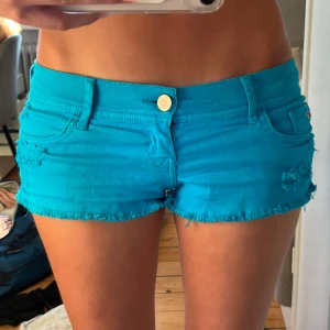 Turkosa jeansshorts från Hollister - Snygga turkosa jeansshorts från Hollister med låg midja och fransade kanter. De har slitna detaljer på fram- och baksidan, klassiska fickor och knappstängning. Perfekta för varma sommardagar och har en cool, avslappnad vibe.