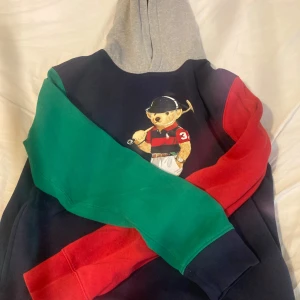Polo Bear hoodie från Ralph Lauren  - Fet hoodie från Polo Ralph Lauren med Polo Bear-tryck på bröstet. Tröjan har grå huva, marinblå kropp, ena ärmen grön och andra röd. Klassisk streetstyle-vibe och riktigt skön bomullskänsla. Perfekt för dig som vill sticka ut lite extra. Det står den är xl men den är för liten för mig som är 170