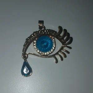 Halsbandshänge i form av öga med blå detalj - Unikt halsbandshänge format som ett öga med långa fransar, dekorerat med glittrande stenar runt en blå iris och en blå droppe som tår. Hängsmycket är i guldtonad metall och har en cool, konstnärlig vibe. PRIS KAN DISKUTERAS!