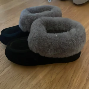Svarta tofflor med grått pälsfoder - Mysiga svarta tofflor med fluffigt grått pälsfoder runt öppningen. Perfekta för att hålla fötterna varma hemma. Sulan är platt och ovandelen är mjuk, vilket gör dem superbekväma att glida i. Stilen är enkel men ändå cozy, och passar dig som gillar chill vibes.