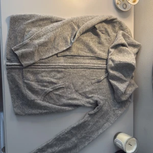 Grå hoodie i kashmir från KSHMR - Mjuk och lyxig grå hoodie från KSHMR i storlek S, tillverkad i kashmir. Tröjan har huva med snörning, dragkedja framtill och ribbade muddar vid ärmslut och nederkant. Perfekt för dig som gillar chill och stilren look.