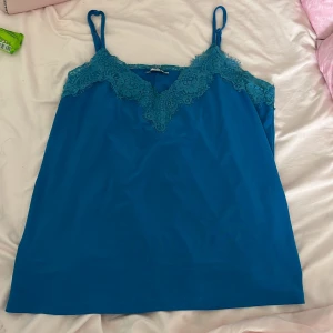 Blå linnetopp med spets från MISU - Supersnygg blå linnetopp från MISU med tunna axelband och v-ringning dekorerad med spetsdetaljer. Perfekt för att ge en chill men ändå stilren vibe till din outfit. Lätt och luftig känsla, passar grymt till jeans eller kjol.