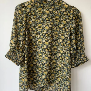 Blommig blus från co' couture - Supersöt blommig blus från co' couture i svart med gula och ljuslila blommor. Blusen har trekvartsärmar med smock vid ärmslut, hög krage och knappar längs ryggen. Tillverkad i tunn polyester som känns lätt och luftig.